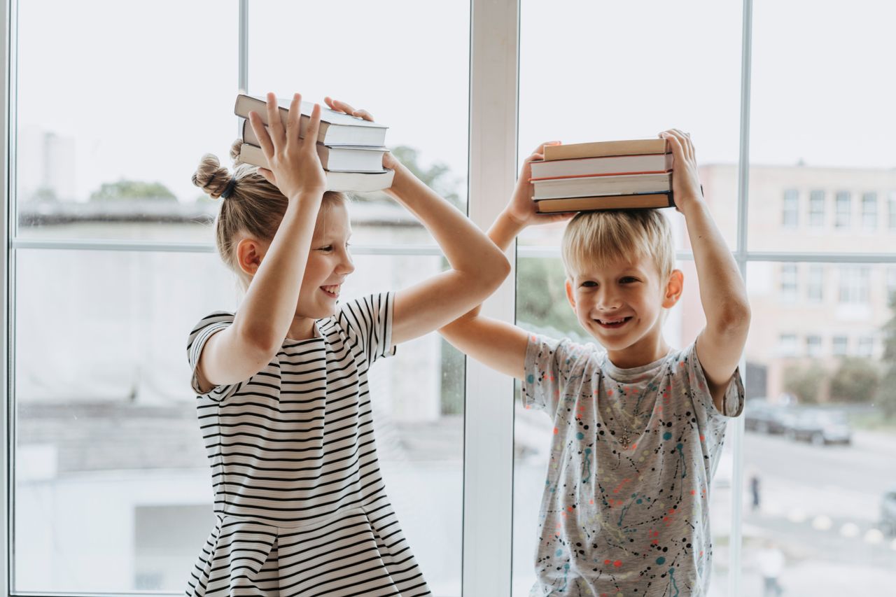 Two laughing siblings playfully balance books on their heads by a window, symbolizing fun learning.