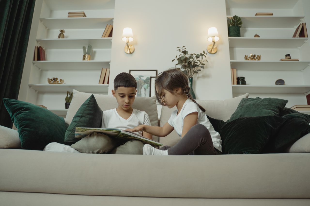 Two children sitting on a sofa reading a book together in a modern living room.
