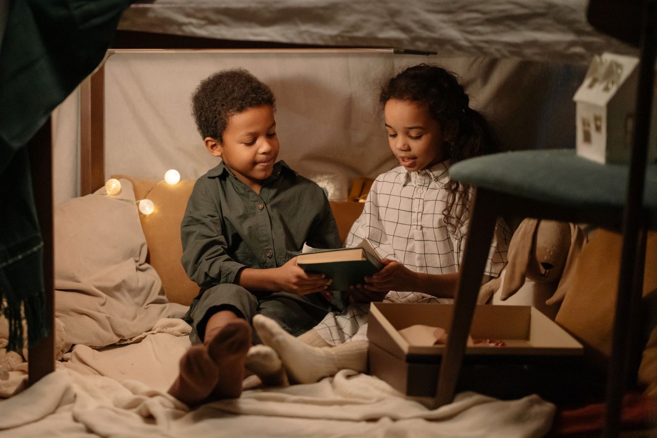 Two children sharing a book in a warm and playful indoor tent setting, fostering sibling bonding.