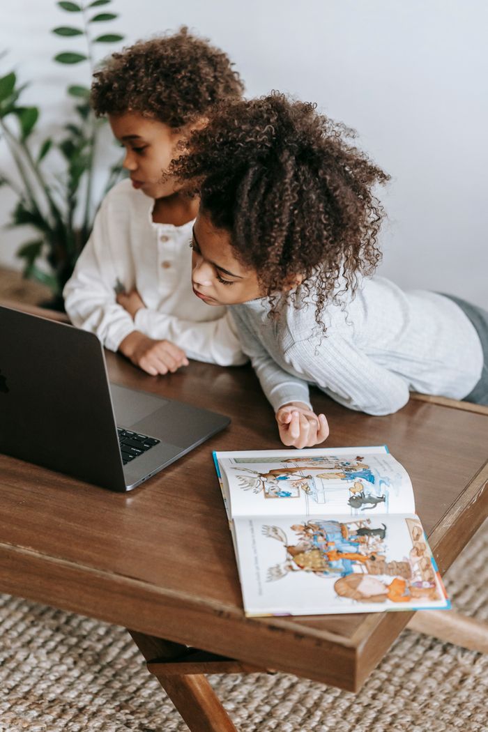 From above of cute little African American girl and boy with curly hair in casual clothes sitting at wooden table and watching video on laptop during weekend at home