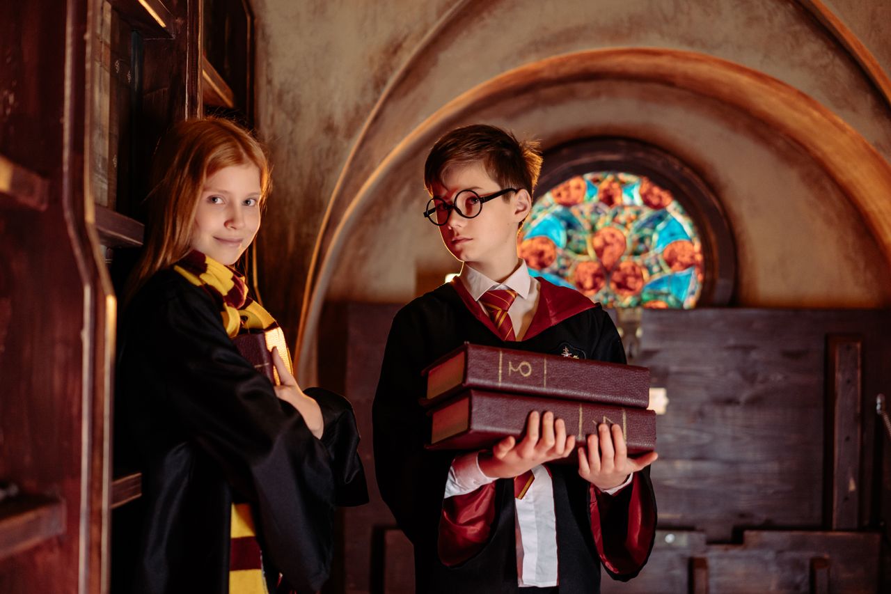 Two children dressed as wizards with robes and glasses, holding books indoors.