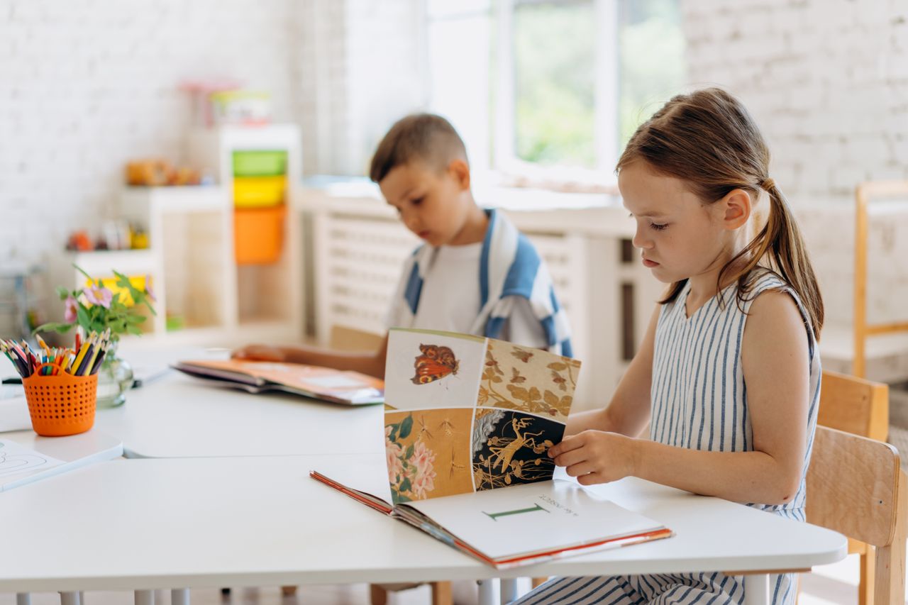 Two children reading books at a bright indoor classroom table filled with colorful supplies.